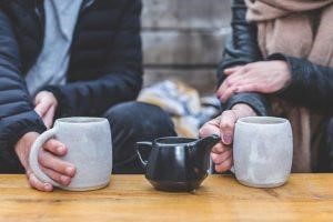Two people holding mugs of tea with a black teapot on a wooden table.